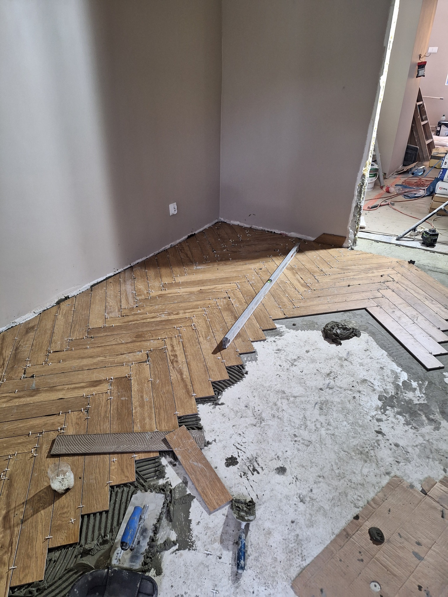 Układanie drewnopodobnej podłogi z płytek w jodełkę, widoczny poziomica, krzyżyki dystansowe i narzędzia glazurnicze.