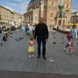 Mężczyzna trzymający za rękę małą dziewczynkę z pluszakiem na tle Rynku Głównego w Krakowie, wokół gołębie