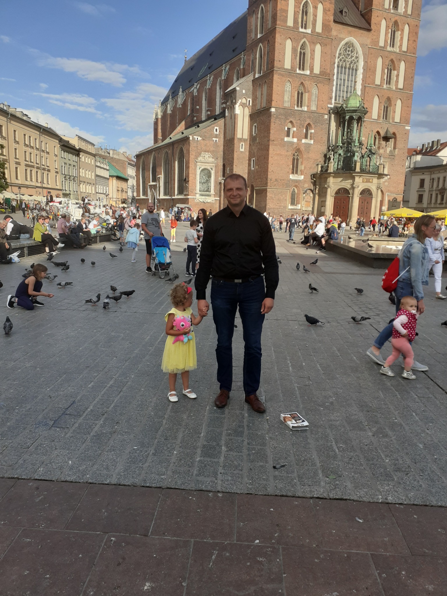 Mężczyzna trzymający za rękę małą dziewczynkę z pluszakiem na tle Rynku Głównego w Krakowie, wokół gołębie