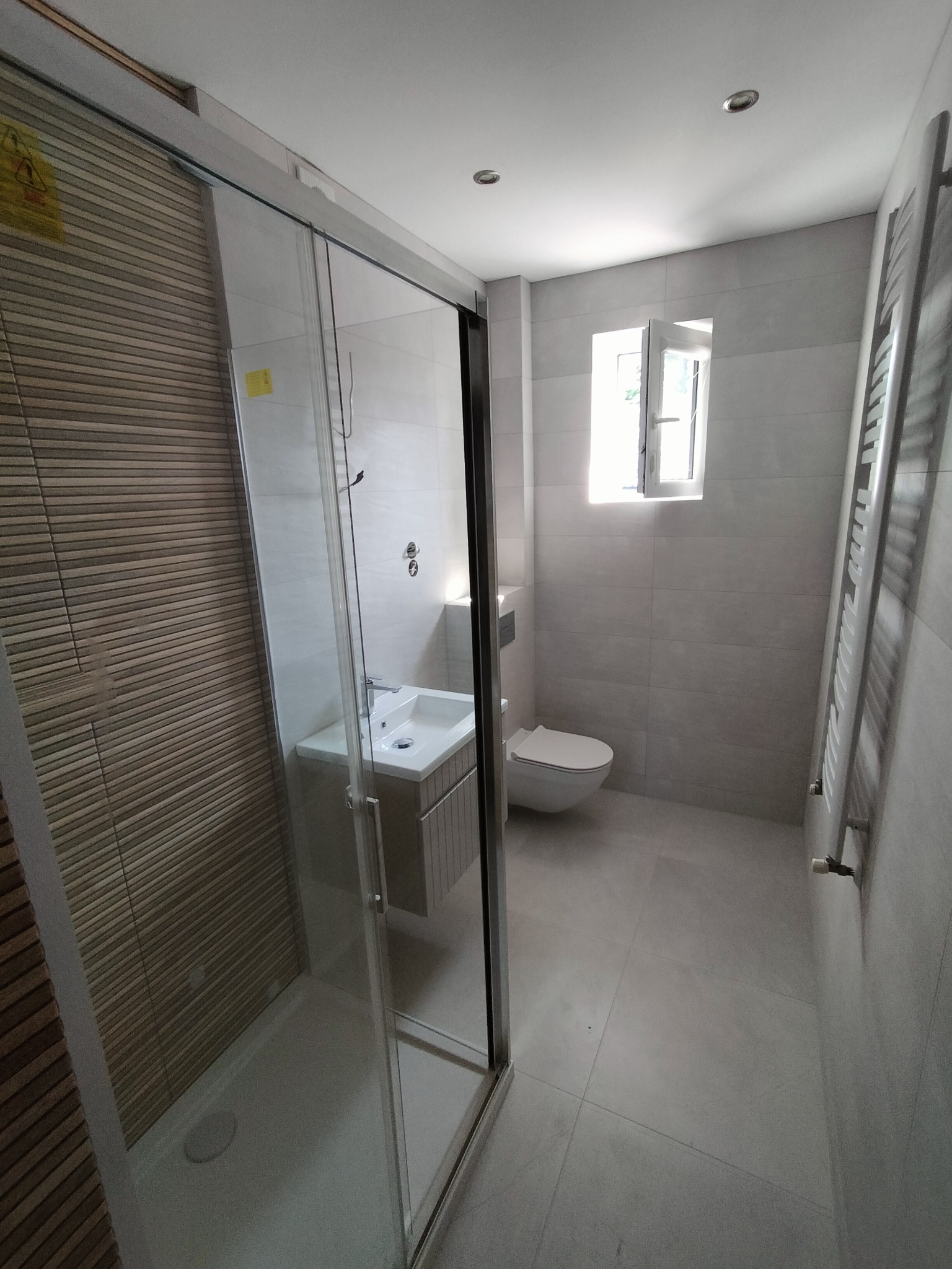 Nowoczesna łazienka z kabiną prysznicową typu walk-in, umywalką z szafką, toaletą i grzejnikiem drabinkowym, utrzymana w szaro-białej kolorystyce z akcentem drewnopodobnych płytek na ścianie...