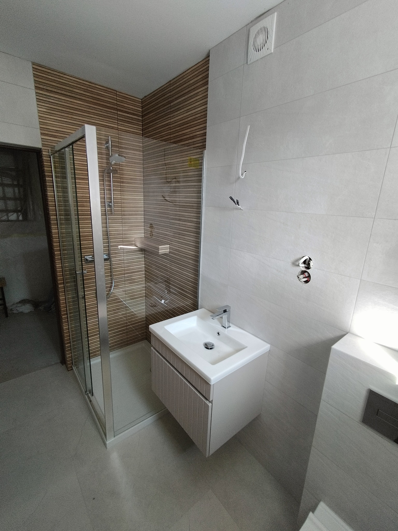 Nowoczesna kabina prysznicowa z deszczownicą i półką, umywalka z szafką w łazience z częściowo ułożonymi płytkami, widoczne niedokończone podłączenia elektryczne.