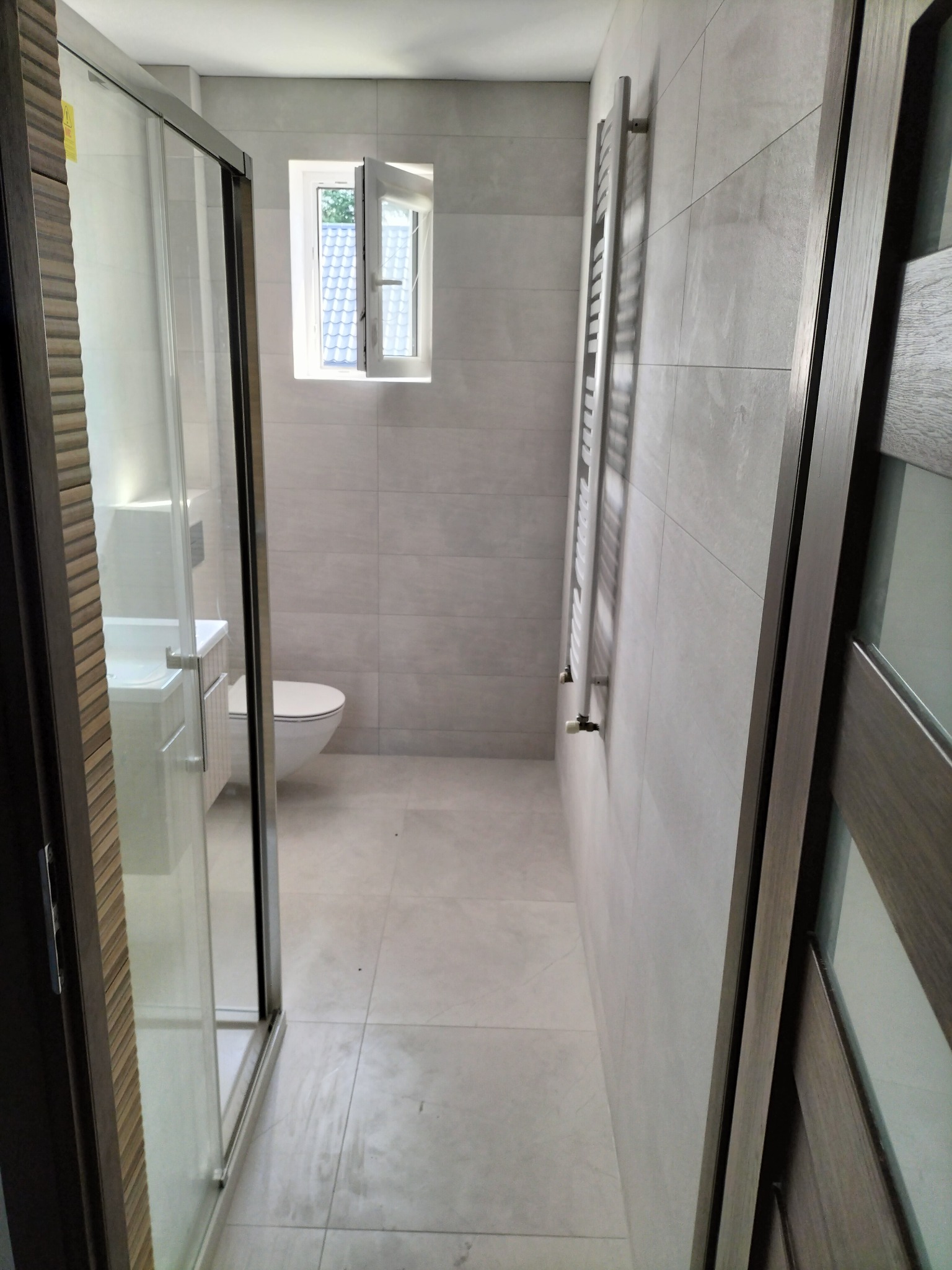 Wykończona łazienka w Wiązownicy z kabiną prysznicową, podwieszaną toaletą i grzejnikiem drabinkowym na ścianie pokrytej szarymi płytkami.