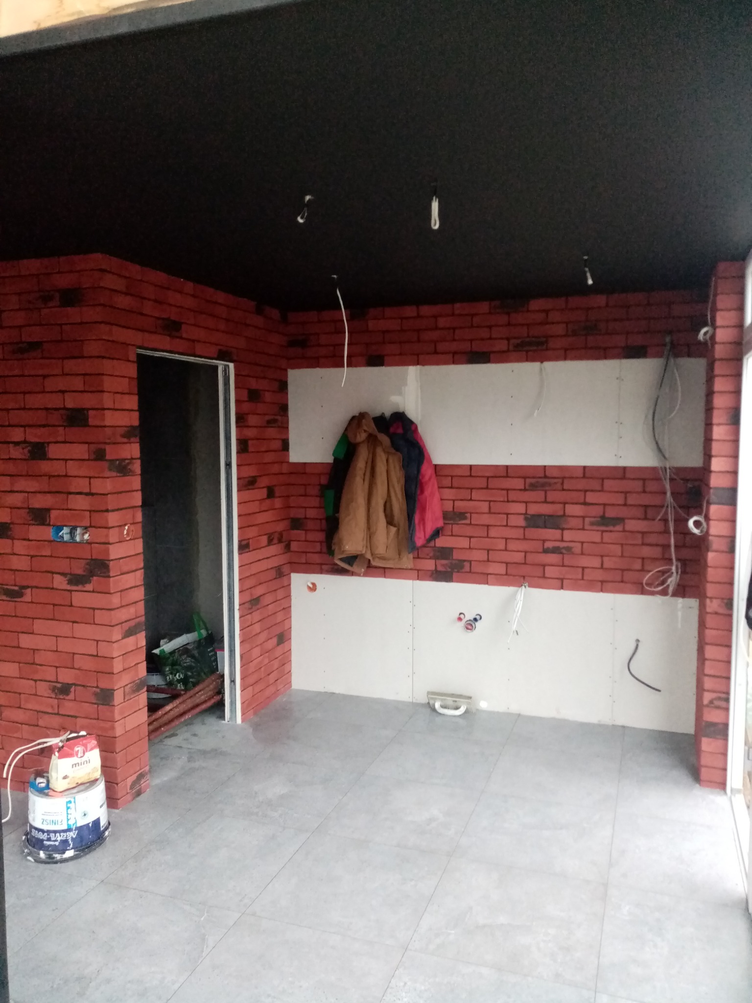 Wnętrze w trakcie prac wykończeniowych: ściany częściowo wyłożone cegłą klinkierową, fragmenty pokryte płytami kartonowo-gipsowymi, widoczne przewody elektryczne, na podłodze szare płytki, przy...