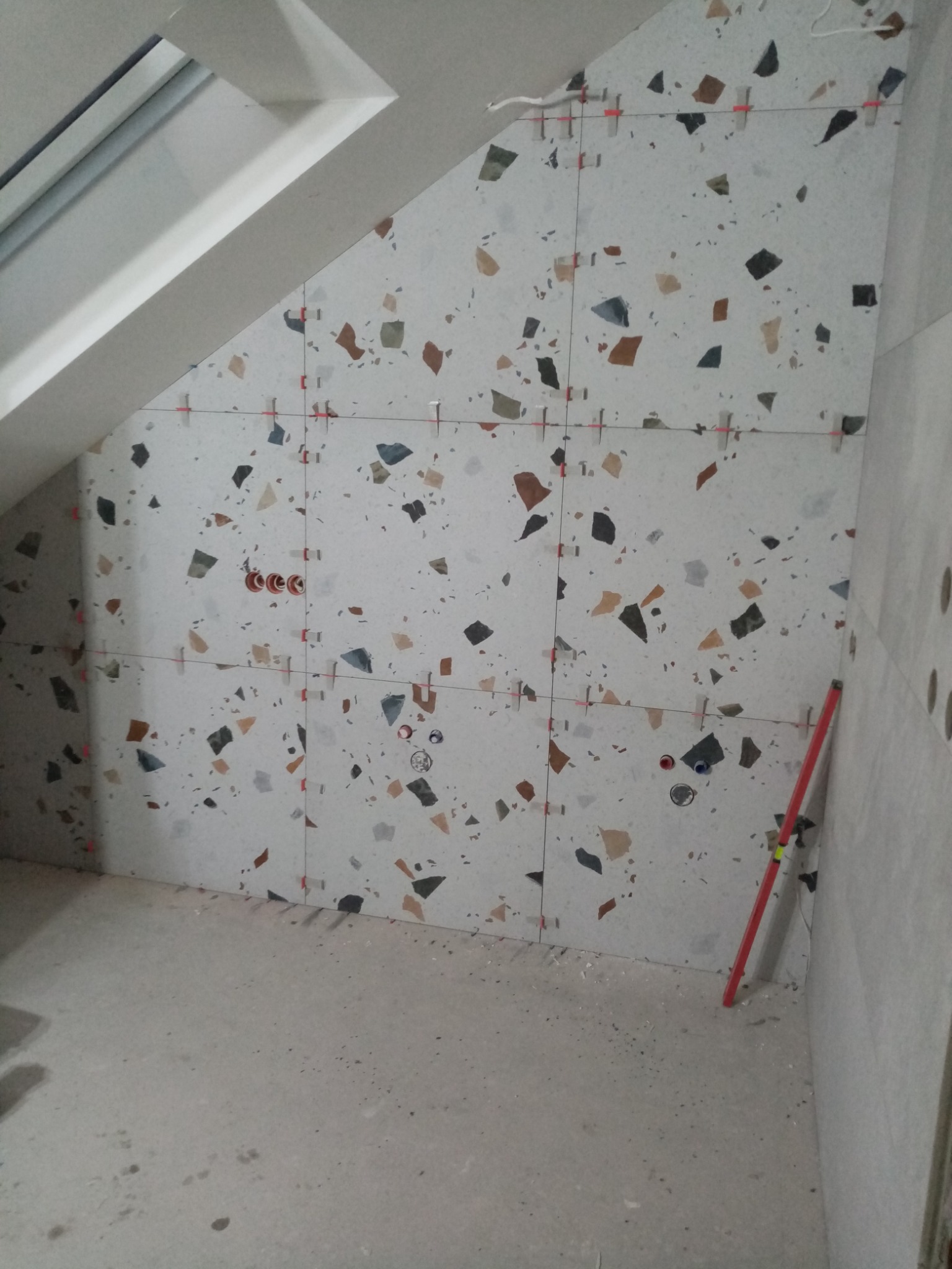 Ściana w łazience na poddaszu, w trakcie układania płytek w stylu lastryko, z widocznymi krzyżykami dystansowymi i poziomnicą przy ścianie.