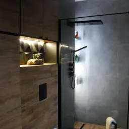 Nowoczesna łazienka z drewnopodobnymi płytkami, kabiną prysznicową typu walk-in z czarnymi ramami i deszczownicą, podświetlaną półką z dekoracją roślinną oraz toaletą.