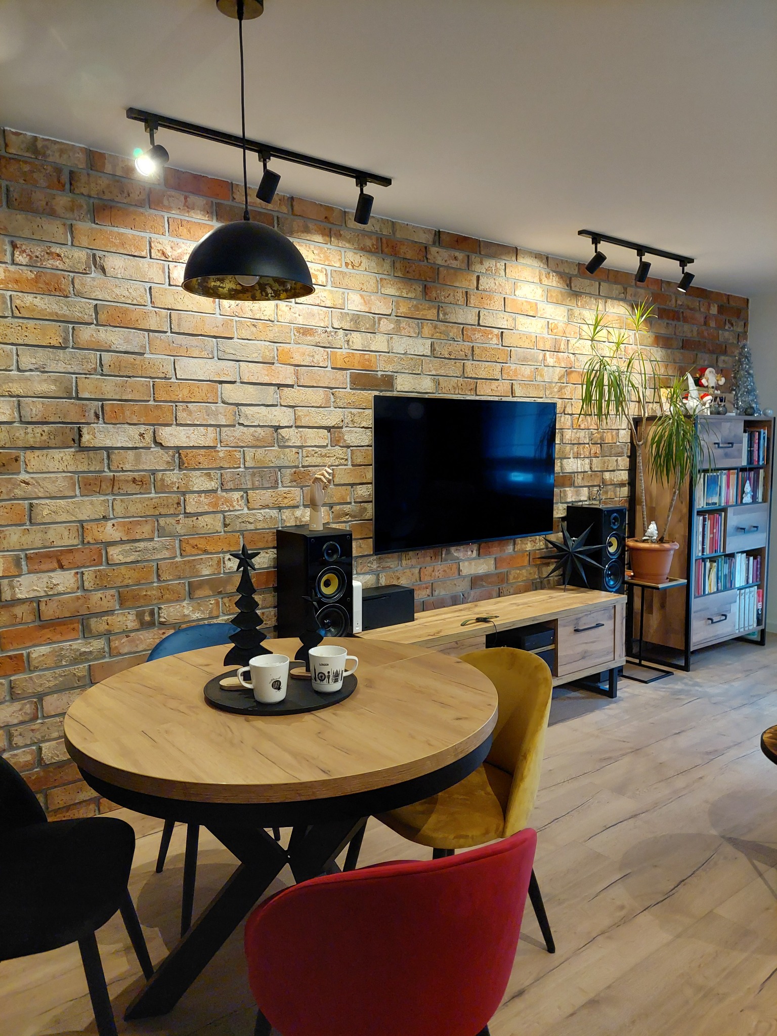 Salon z ceglaną ścianą, oświetleniem punktowym, drewnianym stołem z dekoracjami świątecznymi i kolorowymi krzesłami, w tle szafka RTV i regał z książkami.