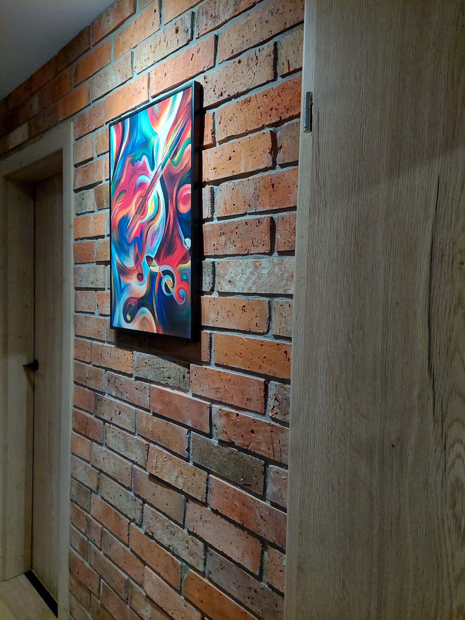Murowana ściana z cegły w Knurowie, ozdobiona abstrakcyjnym obrazem w czarnej ramie przedstawiającym motywy muzyczne, sąsiadująca z drewnianymi drzwiami.