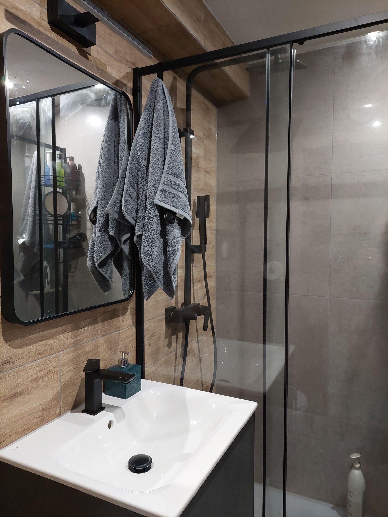 Nowoczesna łazienka z kabiną prysznicową, umywalką nablatową z czarną armaturą i szarymi ręcznikami wiszącymi na czarnej ramie kabiny.