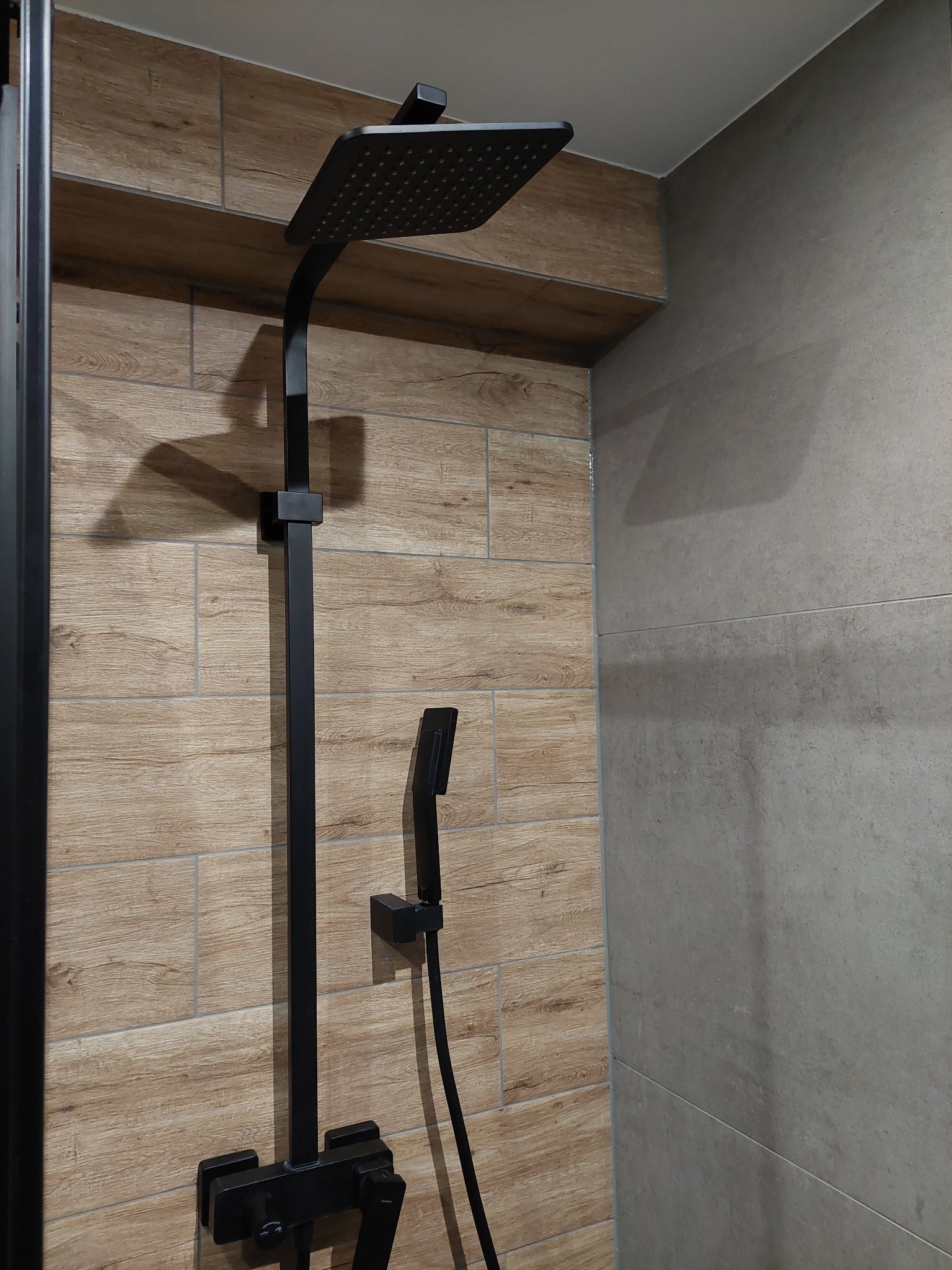 Czarny zestaw prysznicowy z deszczownicą i słuchawką na tle płytek imitujących drewno i szarych płytek w łazience.