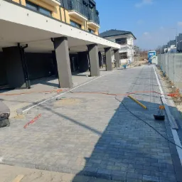 Pracownik w trakcie układania kostki brukowej przed budynkiem wielorodzinnym z kolumnami, widoczne narzędzia, poziomica, sznurek i fragmenty budynku w tle.