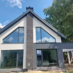 Nowoczesny dom murowany z dużymi oknami, elewacją w odcieniach bieli i szarości oraz ceglanym kominem, widok z dołu na tle błękitnego nieba z chmurami.