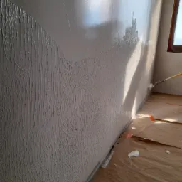 Nakładanie struktury na ścianę w rogu pokoju, widoczne ślady farby na zabezpieczonej podłodze, częściowo wykończona powierzchnia z wyraźną fakturą.