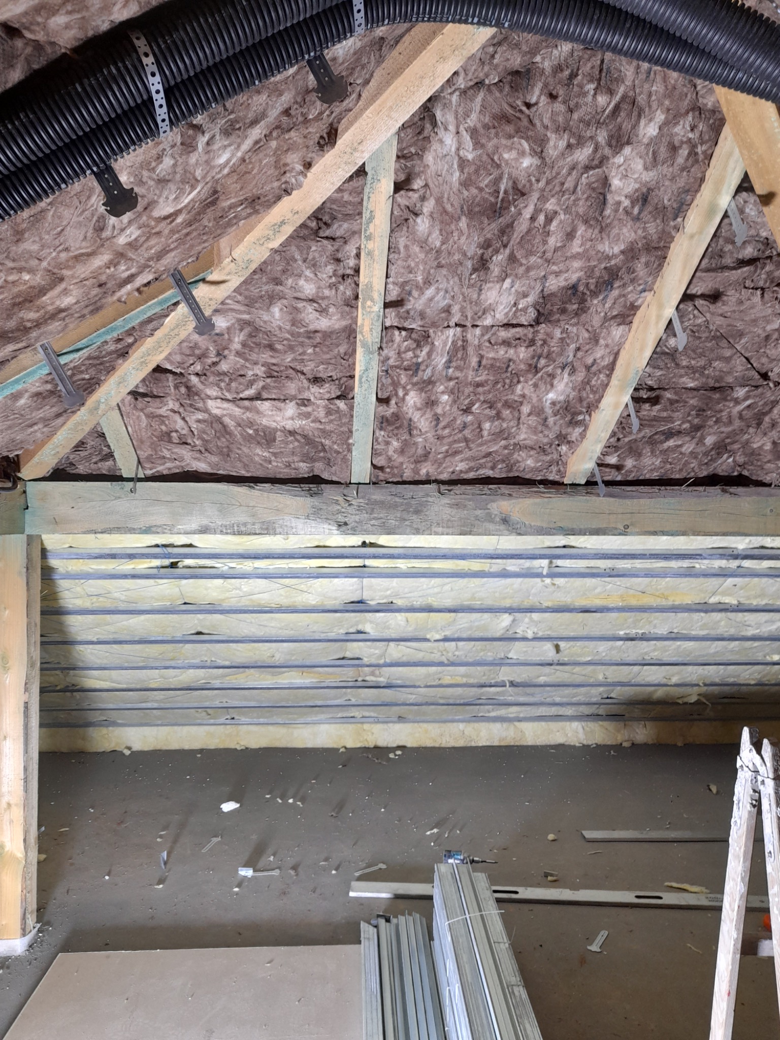 Poddasze w trakcie ocieplania, widoczne krokwie z izolacją z wełny mineralnej, rury wentylacyjne pod sufitem, metalowe profile przy podłodze i drabina oparta o ścianę.