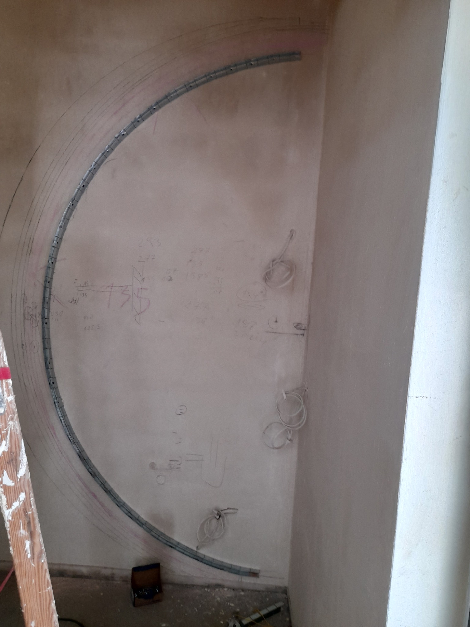 Metalowy profil wygięty w łuk, zamocowany do surowej ściany z naniesionymi szkicami i pomiarami, przygotowanie do montażu konstrukcji.