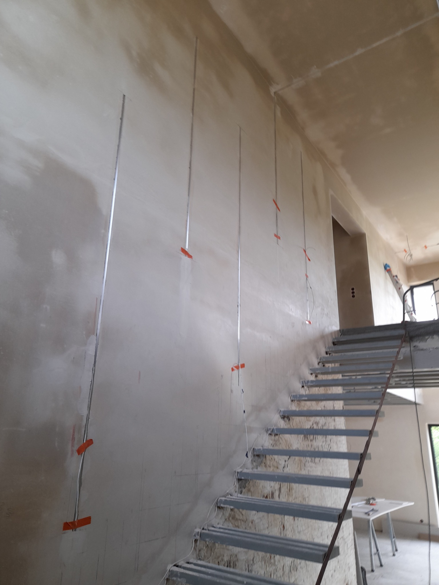 Ściana po nałożeniu pierwszej warstwy gładzi, z pionowo poprowadzonymi przewodami elektrycznymi przytwierdzonymi pomarańczową taśmą, widoczne metalowe schody w surowym stanie i zarysowane linie...