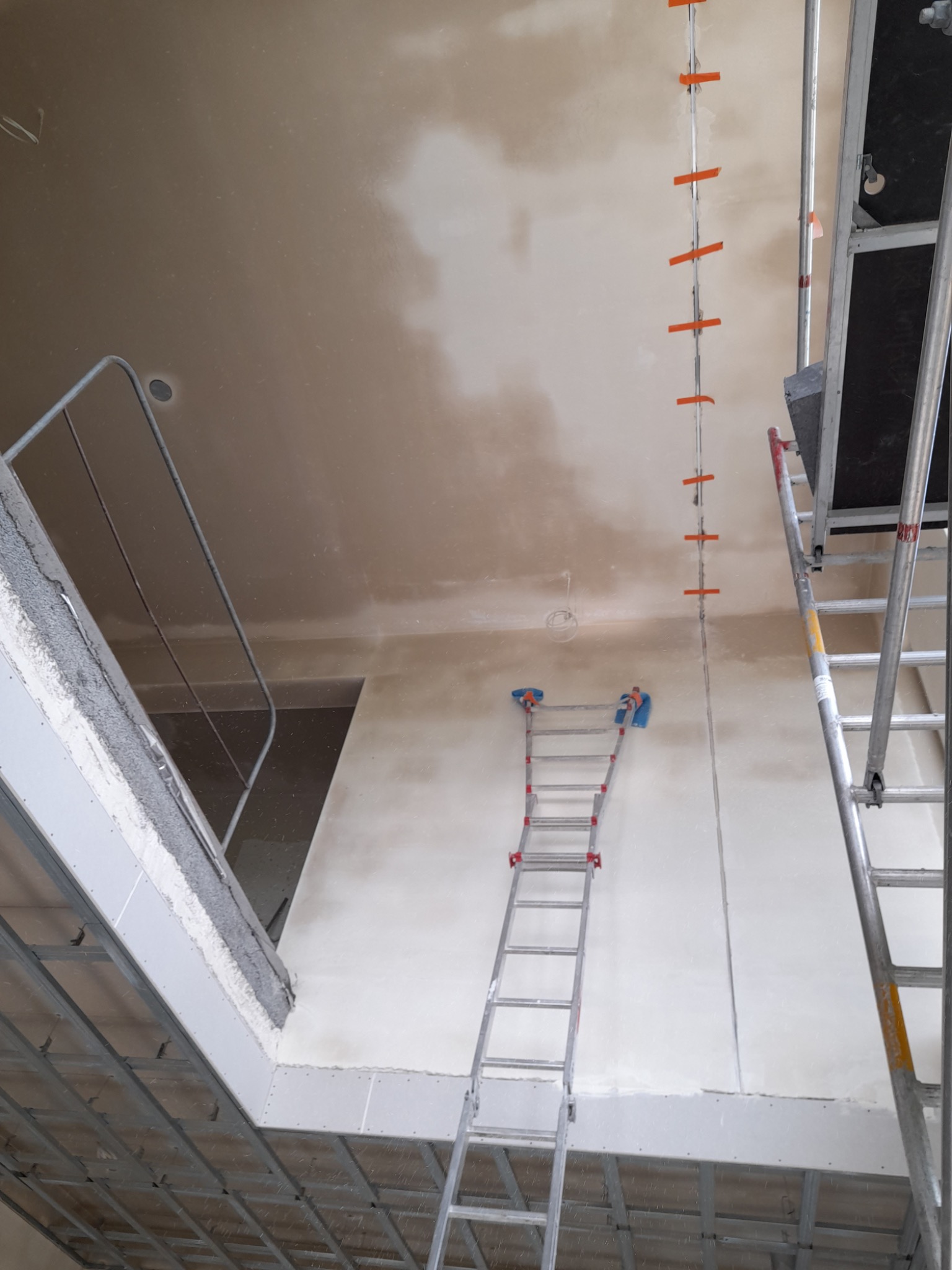Wnętrze w trakcie prac wykończeniowych: ściany przygotowane do malowania, widoczne drabiny, metalowa konstrukcja sufitu podwieszanego i pionowa listwa z pomarańczowymi znacznikami.