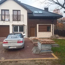 Dom dwukondygnacyjny z elementami w trakcie wykończenia: okno zabezpieczone folią, obok zaparkowany samochód z reklamą firmy remontowej, na podjeździe ułożone palety z kostką brukową.