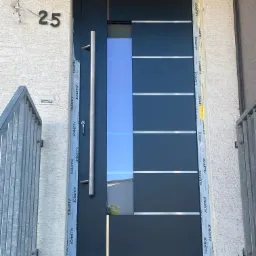 Nowoczesne, antracytowe drzwi wejściowe z przeszkleniem i długim, pionowym uchwytem ze stali nierdzewnej, zamontowane w elewacji z numerem domu 25, z widocznymi taśmami ochronnymi producenta...