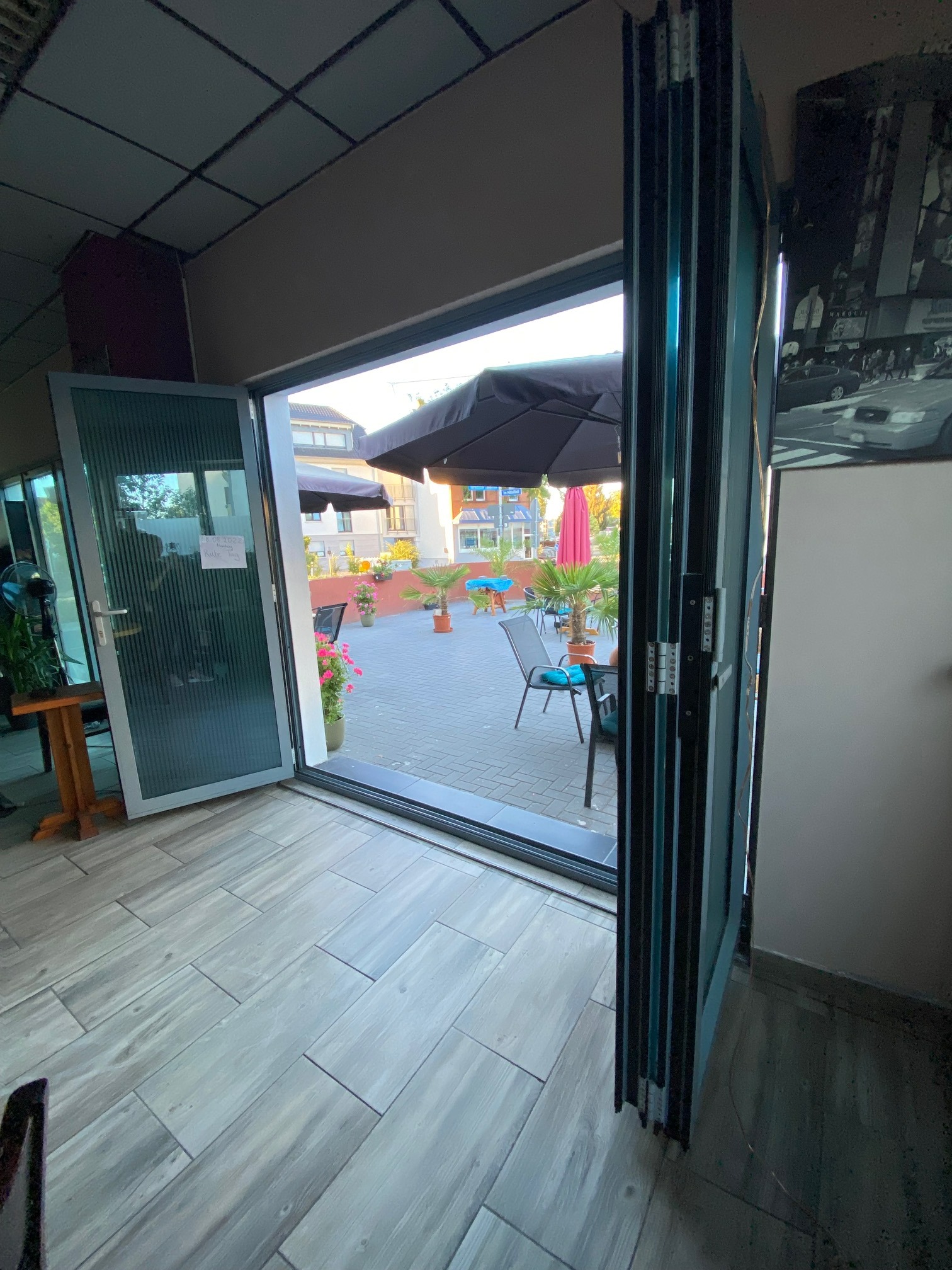 Otwarta, składana aluminiowa ściana drzwiowa harmonijkowa w kolorze RAL 7001, z widokiem na patio z parasolami i kwiatami, zamontowana w lokalu usługowym z szarą podłogą imitującą drewno.