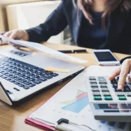 Kobieta w czarnej marynarce przy biurku, obliczenia na kalkulatorze, laptop, smartfon, dokumenty z wykresami, miękkie, naturalne światło z lewej strony.