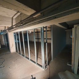 Usługi remontowe - Metalowa konstrukcja szkieletowa ścianki działowej z płyt kartonowo-gipsowych w surowym, betonowym wnętrzu piwnicy, widoczne przewody pod sufitem i fragment sylwetki robotnika.