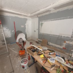 Usługi remontowe - Pracownik przygotowuje masę szpachlową w pomarańczowym wiadrze, w tle pokój w trakcie prac wykończeniowych: częściowo zagruntowane ściany na szaro, widoczny stół z narzędziami, drabina, puszka...