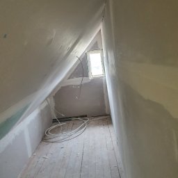 Usługi remontowe - Poddasze w trakcie prac wykończeniowych: ściany pokryte gładzią gipsową, widoczne przewody elektryczne, fragment wełny mineralnej na podłodze i małe okno.