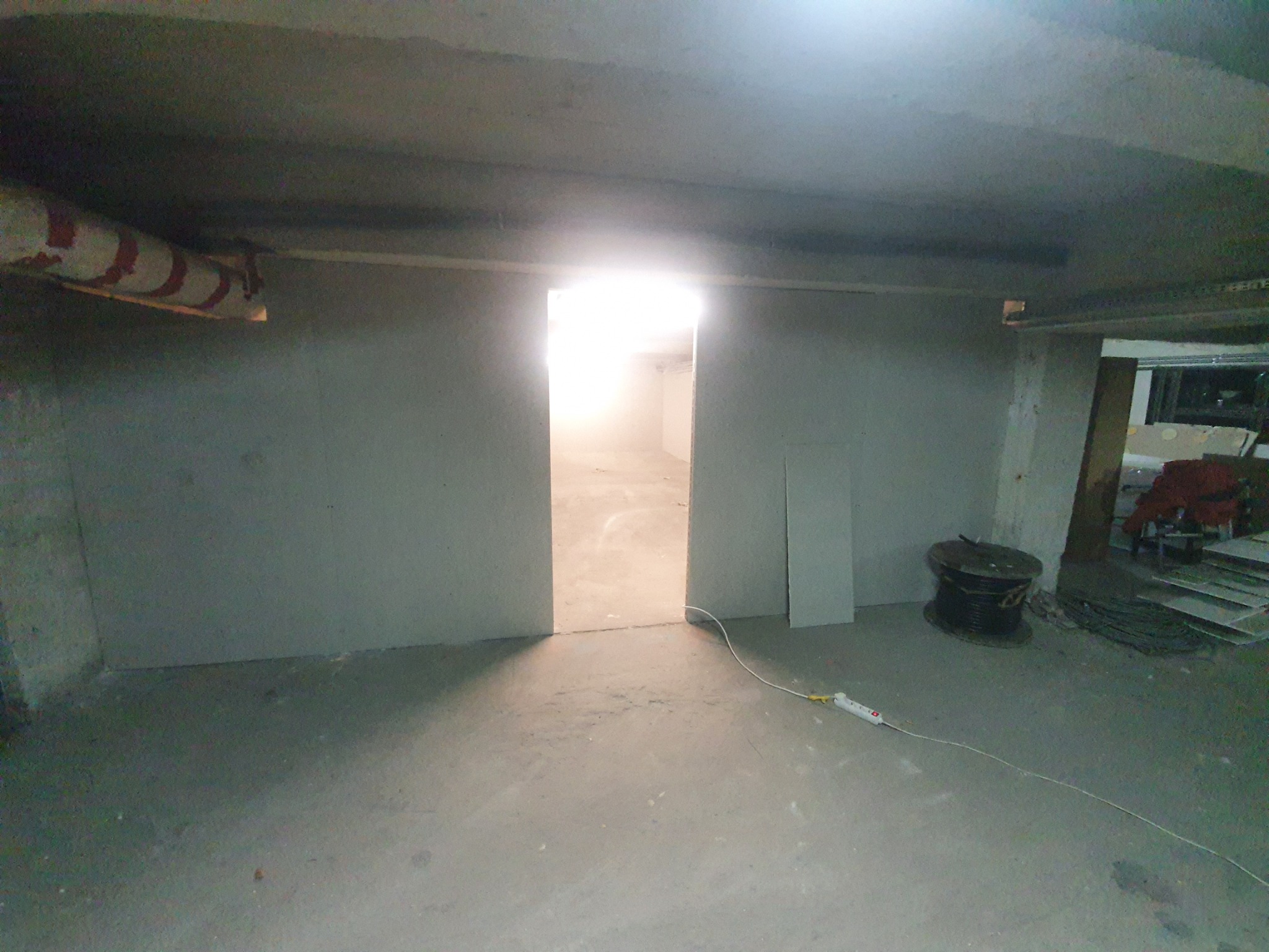Wnętrze w trakcie budowy: surowy betonowy sufit, ściany z płyt gipsowo-kartonowych tworzące przejście do jasnego pomieszczenia, zwój kabla elektrycznego i przedłużacz na podłodze.