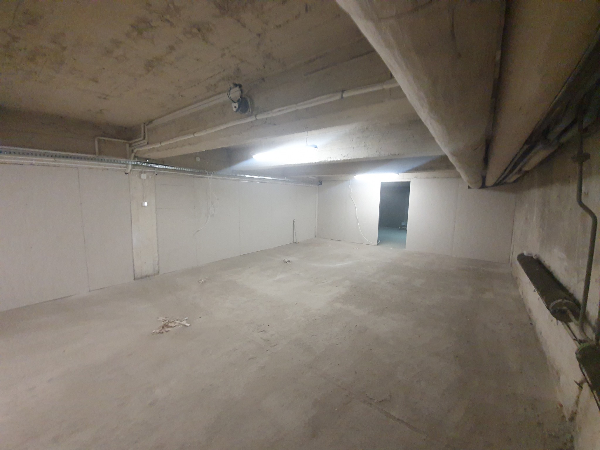 Surowe wnętrze piwnicy z betonową podłogą i sufitem, częściowo wykończone ścianami z płyt kartonowo-gipsowych, widoczne rury i instalacje elektryczne, surowe oświetlenie.