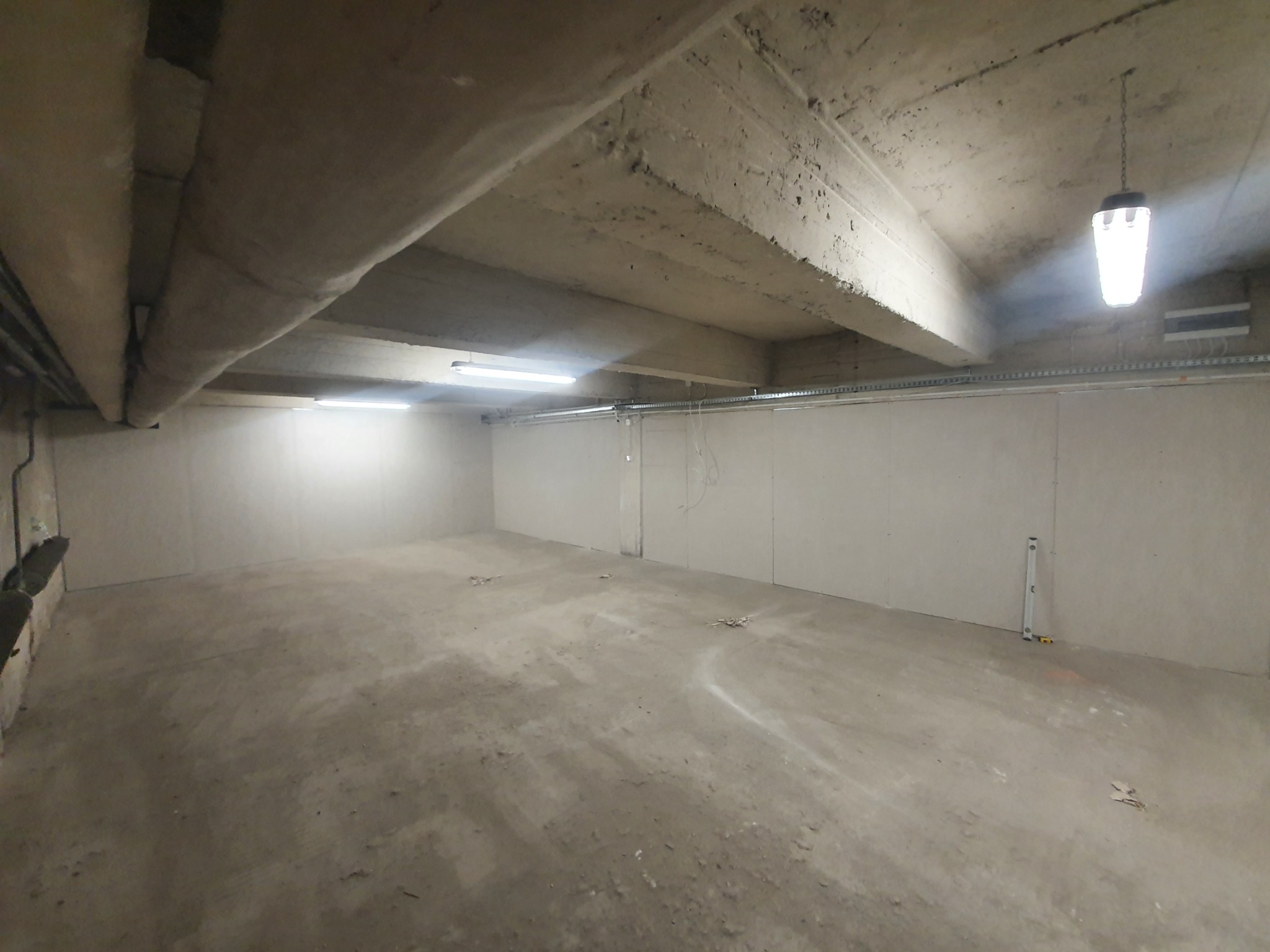 Surowe wnętrze piwnicy z betonowym sufitem i ścianami wyłożonymi płytami gipsowo-kartonowymi, wisząca lampa przemysłowa, widoczny poziomica przy ścianie.