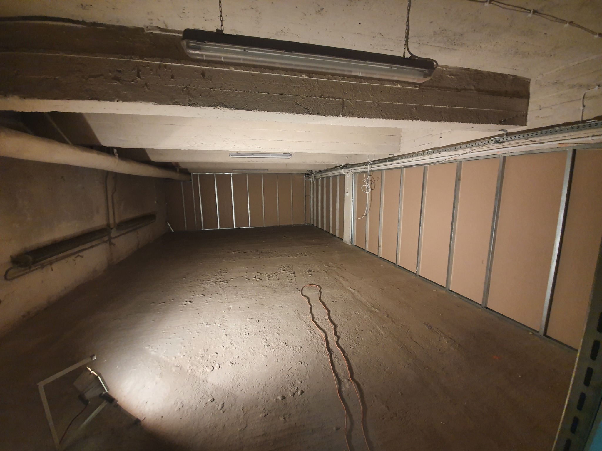 Poddasze w trakcie adaptacji: surowy betonowy strop, metalowe profile i płyty kartonowo-gipsowe, widoczna lampa budowlana i kabel na podłodze.