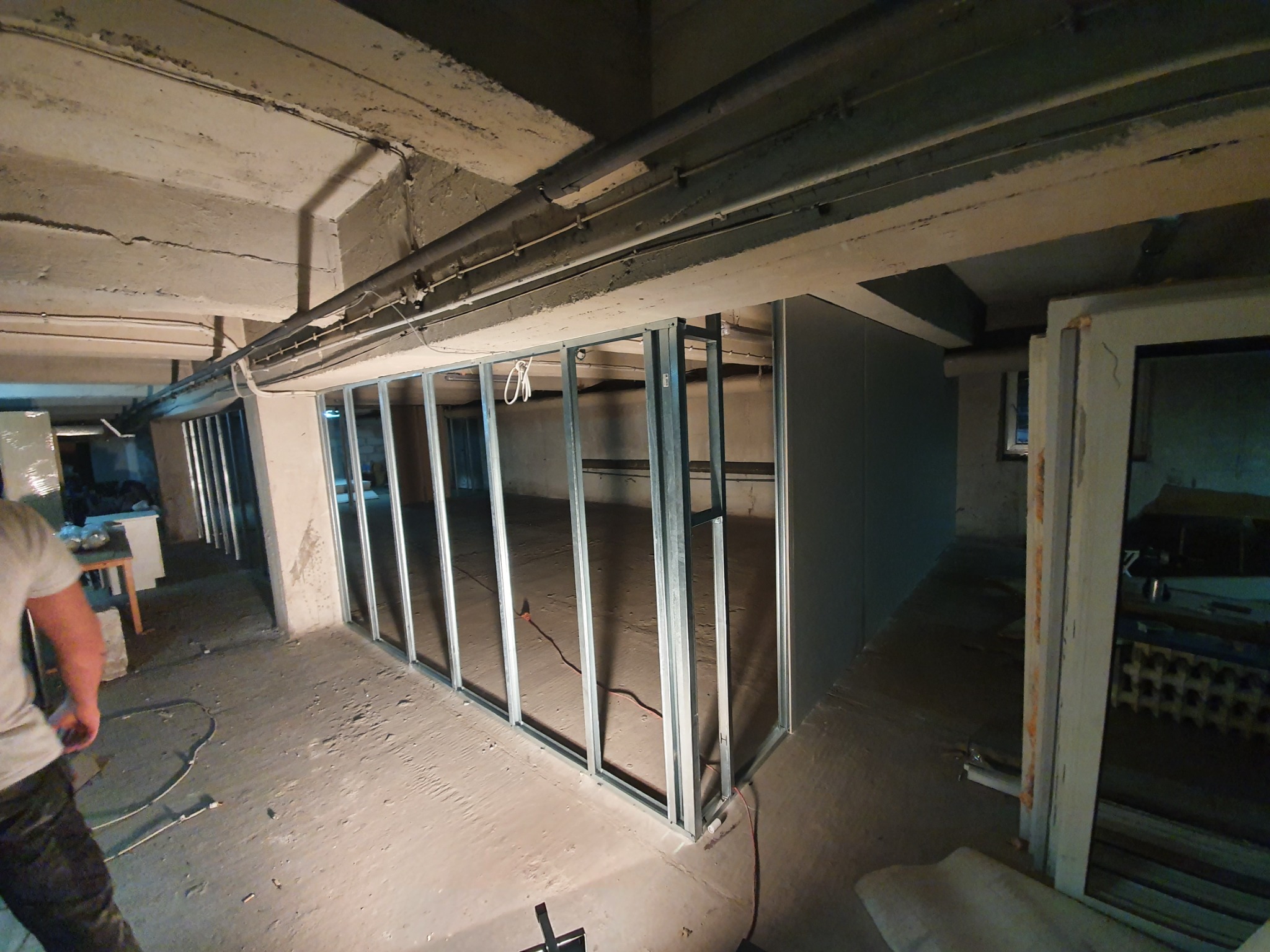 Metalowa konstrukcja szkieletowa ścianki działowej z płyt kartonowo-gipsowych w surowym, betonowym wnętrzu piwnicy, widoczne przewody pod sufitem i fragment sylwetki robotnika.