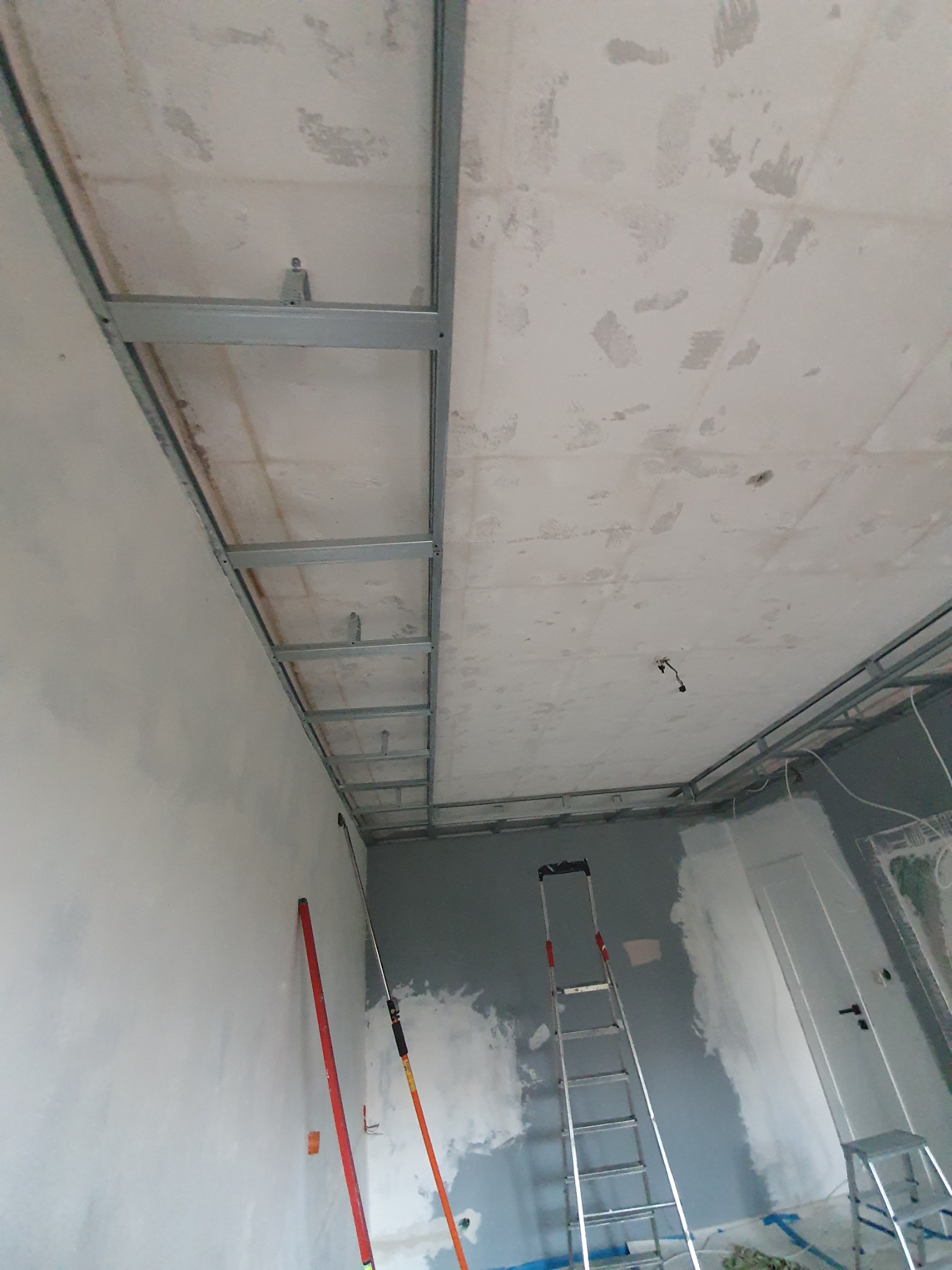 Stelaż metalowy pod konstrukcję sufitu podwieszanego w trakcie montażu, widoczna drabina i narzędzia malarskie przy ścianie, surowy stan wykończenia wnętrza.