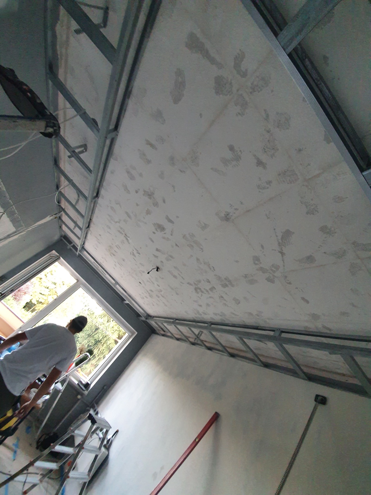 Montaż metalowej konstrukcji sufitu podwieszanego z widocznymi śladami szpachlowania na płycie, drabina i narzędzia w tle, mężczyzna przy oknie.