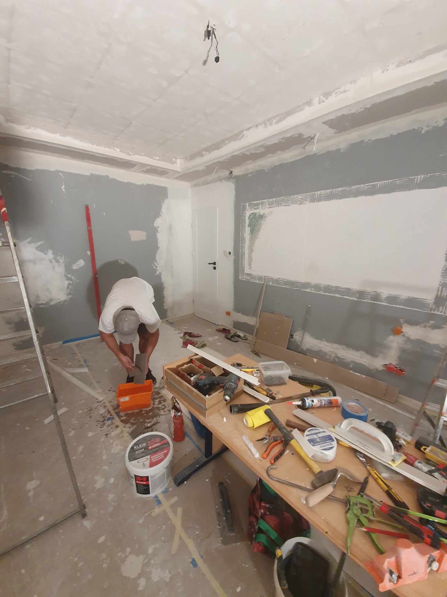 Pracownik przygotowuje masę szpachlową w pomarańczowym wiadrze, w tle pokój w trakcie prac wykończeniowych: częściowo zagruntowane ściany na szaro, widoczny stół z narzędziami, drabina, puszka...