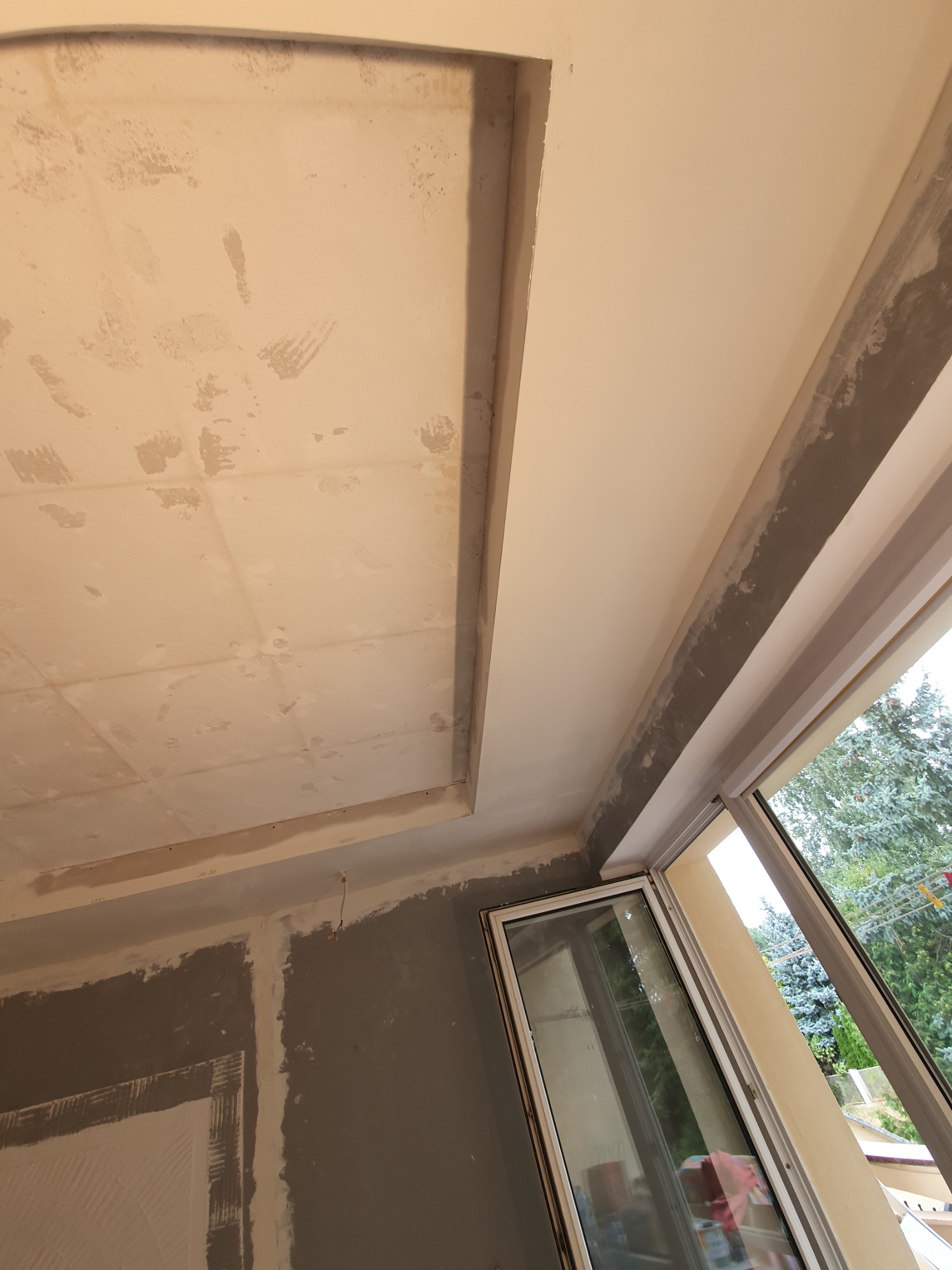 Niedokończony sufit z płyt kartonowo-gipsowych z widocznymi śladami szpachlowania, surowa ściana i okno z widokiem na drzewa.