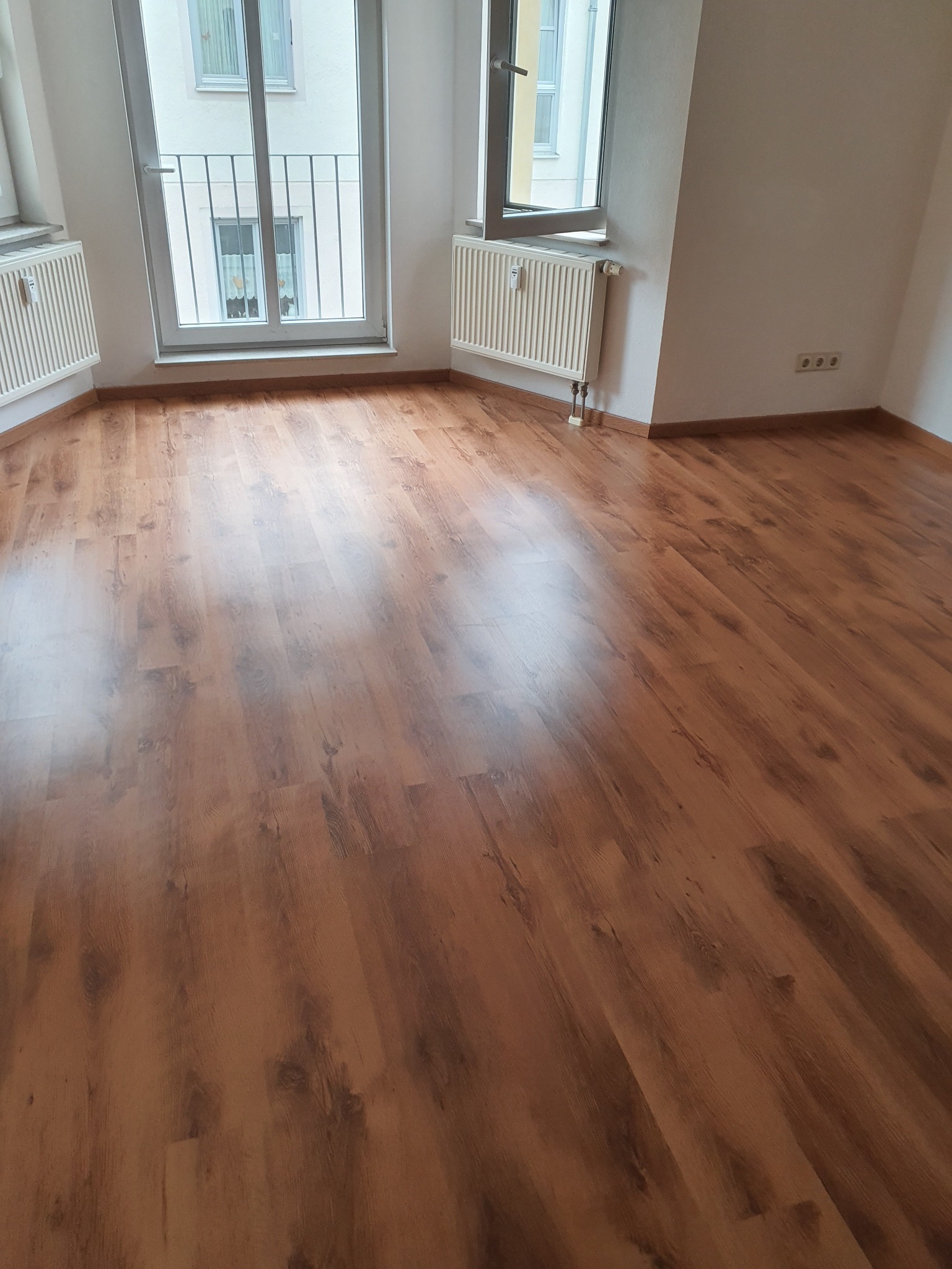 Nowo ułożona, błyszcząca podłoga z paneli w pustym pokoju z białymi ścianami, grzejnikami i oknem balkonowym z metalową barierką.