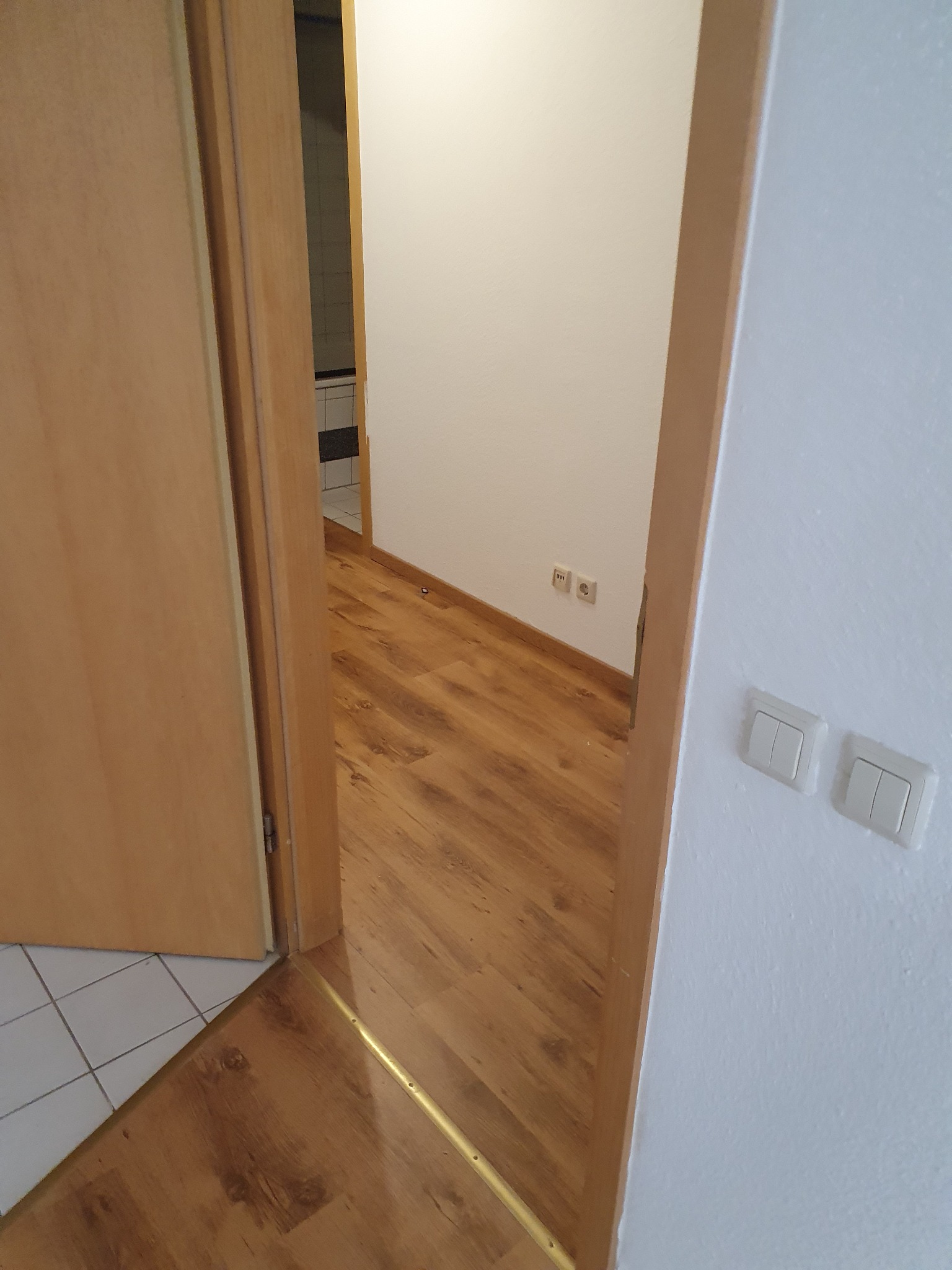 Widok z korytarza na pokój z drewnianą podłogą i białą ścianą, widoczne gniazdka elektryczne i fragment łazienki z białymi płytkami w tle.