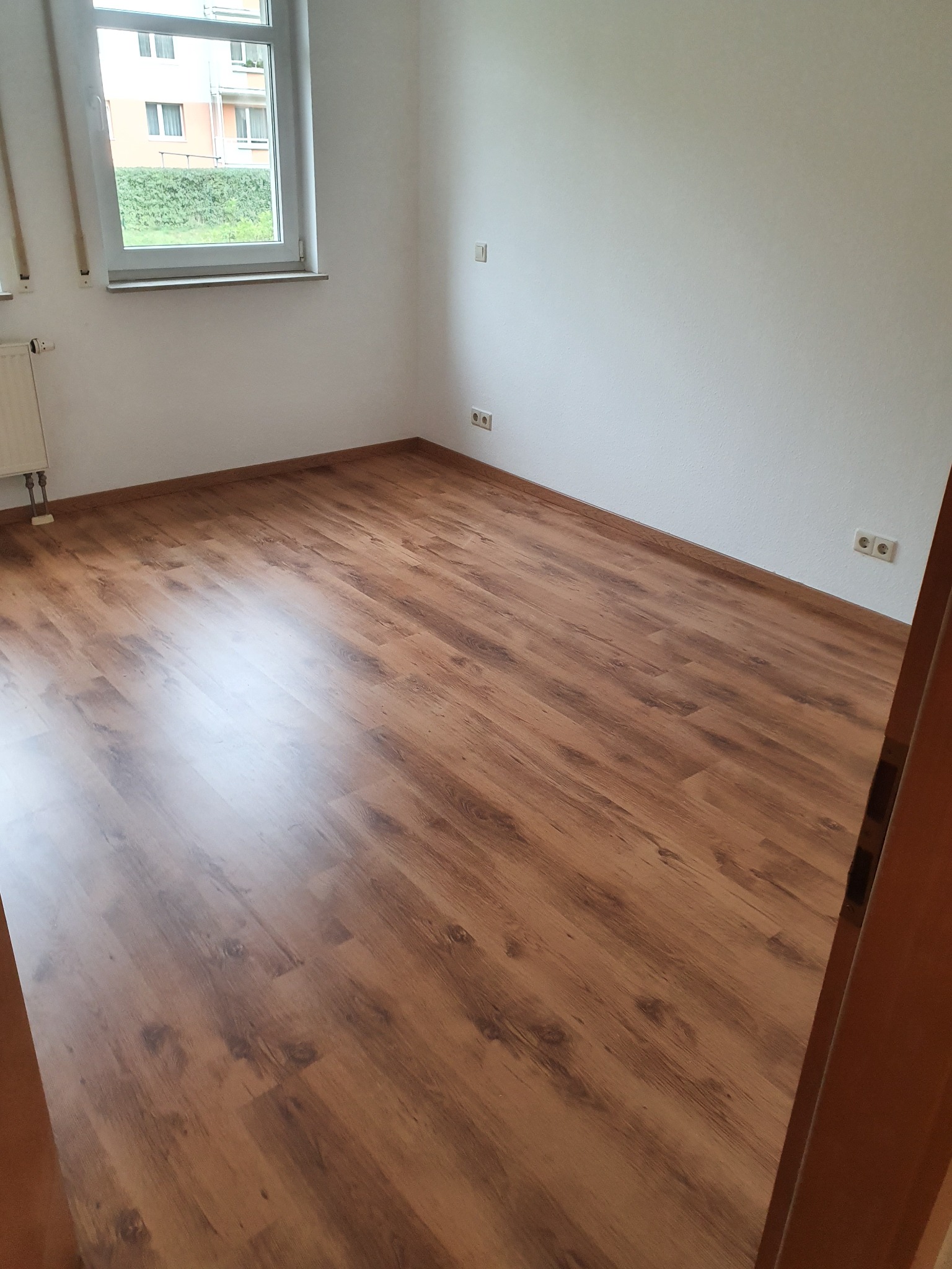 Pusta, jasna sypialnia z panelami podłogowymi w odcieniach brązu, widok na okno z zielenią na zewnątrz i białe ściany z gniazdkami elektrycznymi.