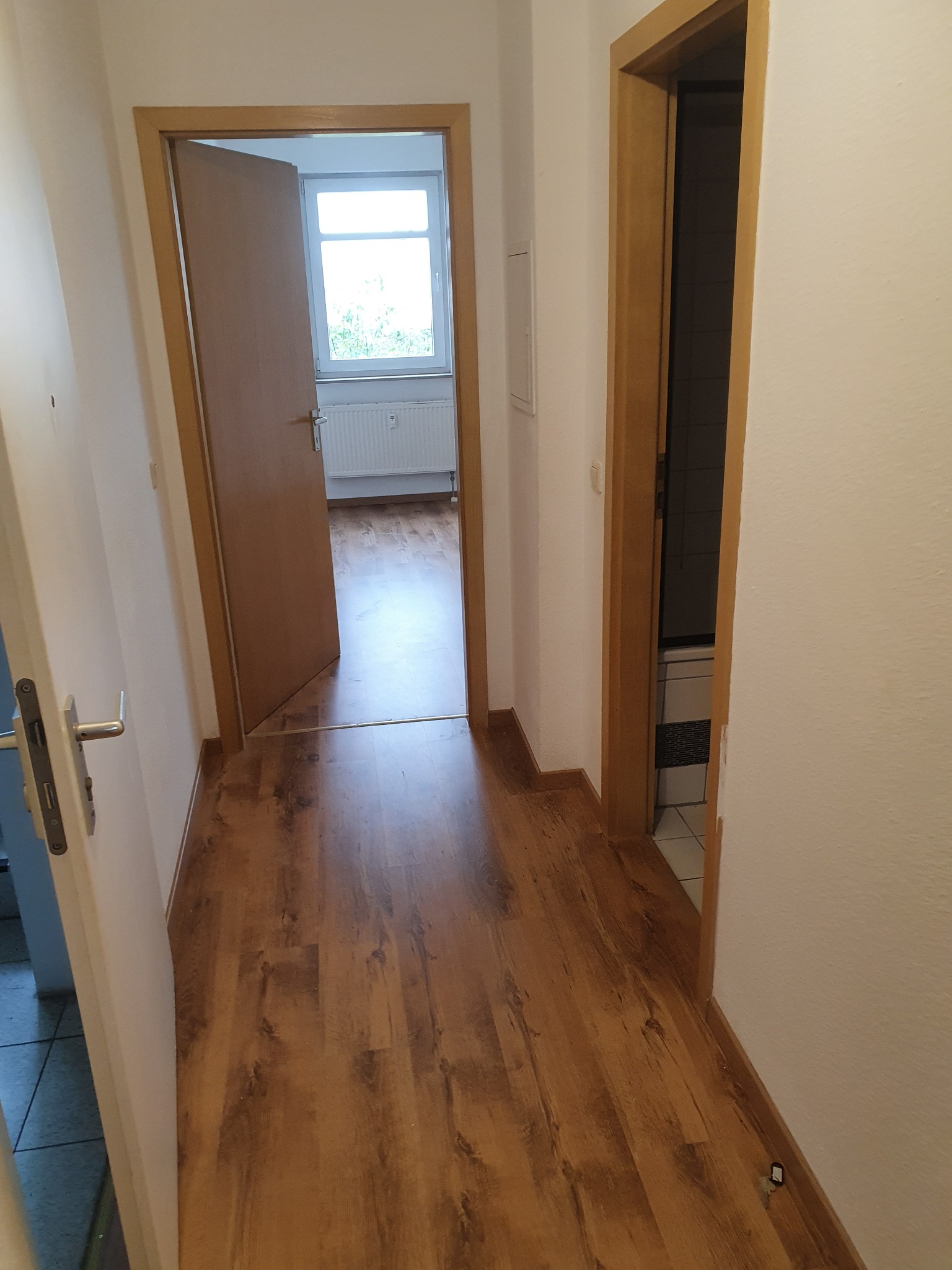 Korytarz z panelami podłogowymi w odcieniach brązu, widok na otwarte drzwi do pokoju z oknem i grzejnikiem oraz wejście do łazienki.