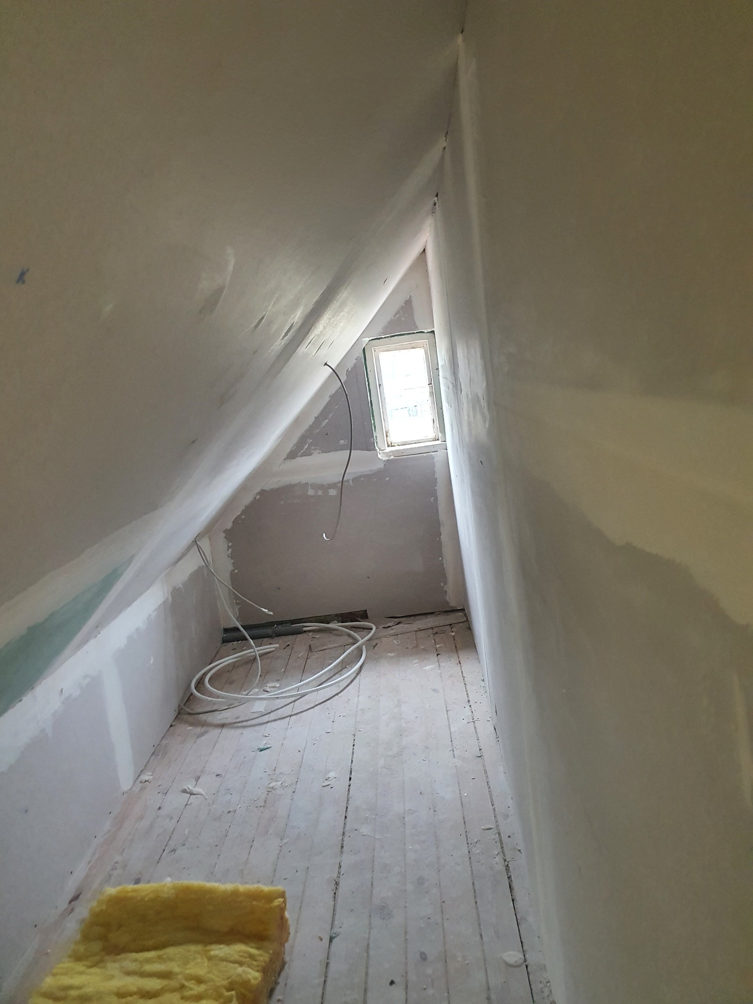 Poddasze w trakcie prac wykończeniowych: ściany pokryte gładzią gipsową, widoczne przewody elektryczne, fragment wełny mineralnej na podłodze i małe okno.