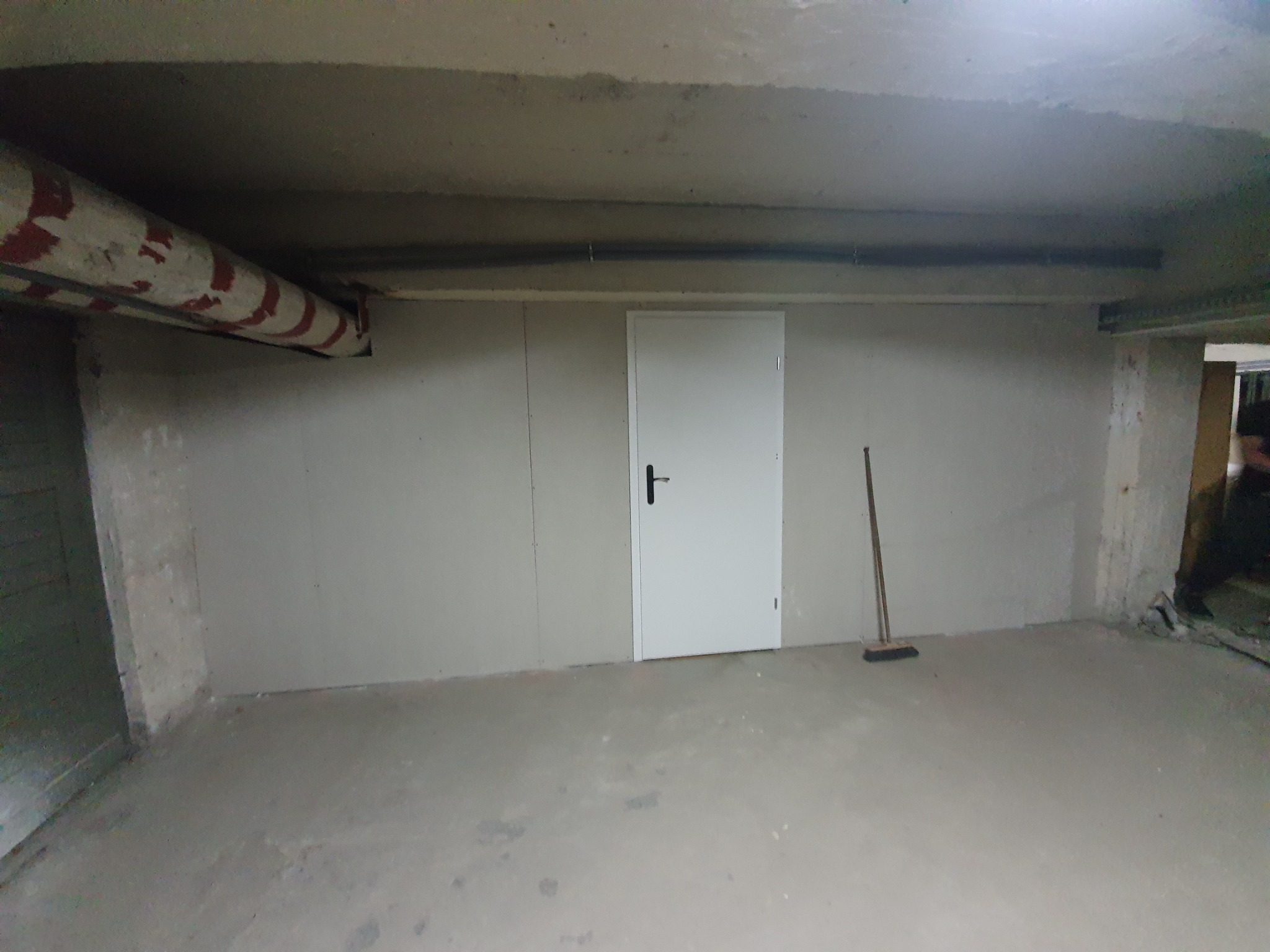 Wykończone pomieszczenie w piwnicy z płytami kartonowo-gipsowymi na ścianach, białe drzwi i betonowa podłoga, rura z czerwonymi paskami pod sufitem, widoczna szczotka na podłodze.