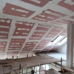 Poddasze w trakcie prac wykończeniowych: widoczne płyty kartonowo-gipsowe na skosach dachu, spoiny wypełnione masą szpachlową, drewniany pomost roboczy, okno dachowe i fragment surowej ściany...