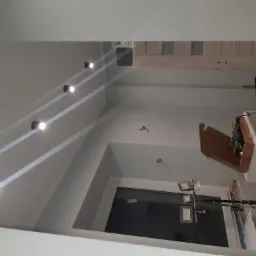 Nowoczesne oświetlenie sufitowe z reflektorami punktowymi w trakcie montażu, widoczne narzędzia i oświetlenie robocze, surowy stan wykończenia wnętrza.