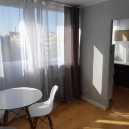 Jasne wnętrze mieszkania z widokiem na budynek przez okno z białymi i szarymi zasłonami, biały okrągły stół i krzesła na drewnianej podłodze, widok na kuchnię w tle.