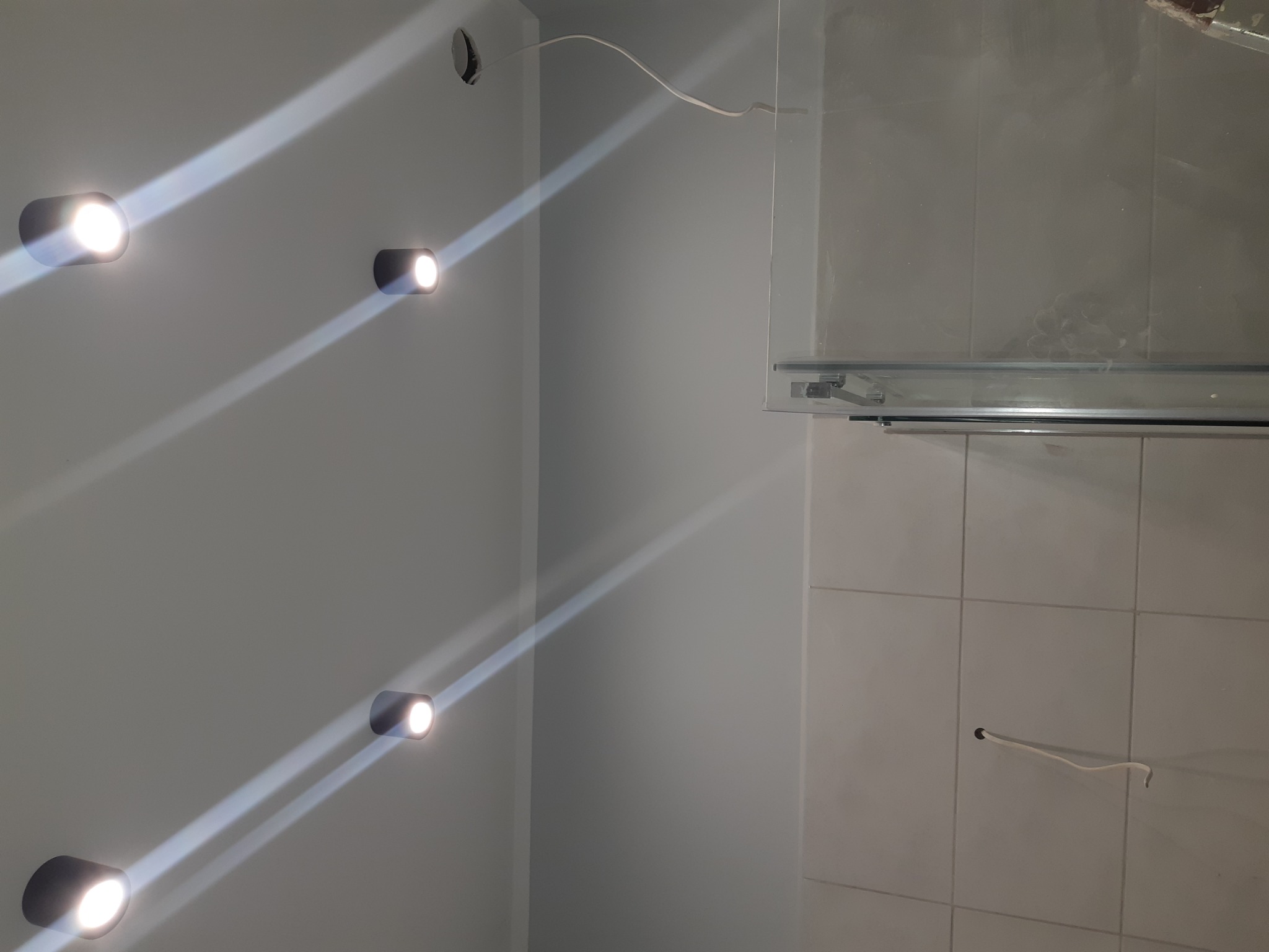 Świeżo zamontowane, okrągłe lampy LED w rzędzie na szarej ścianie, z widocznymi przewodami wystającymi ze ściany w łazience z szarą glazurą i kabiną prysznicową.