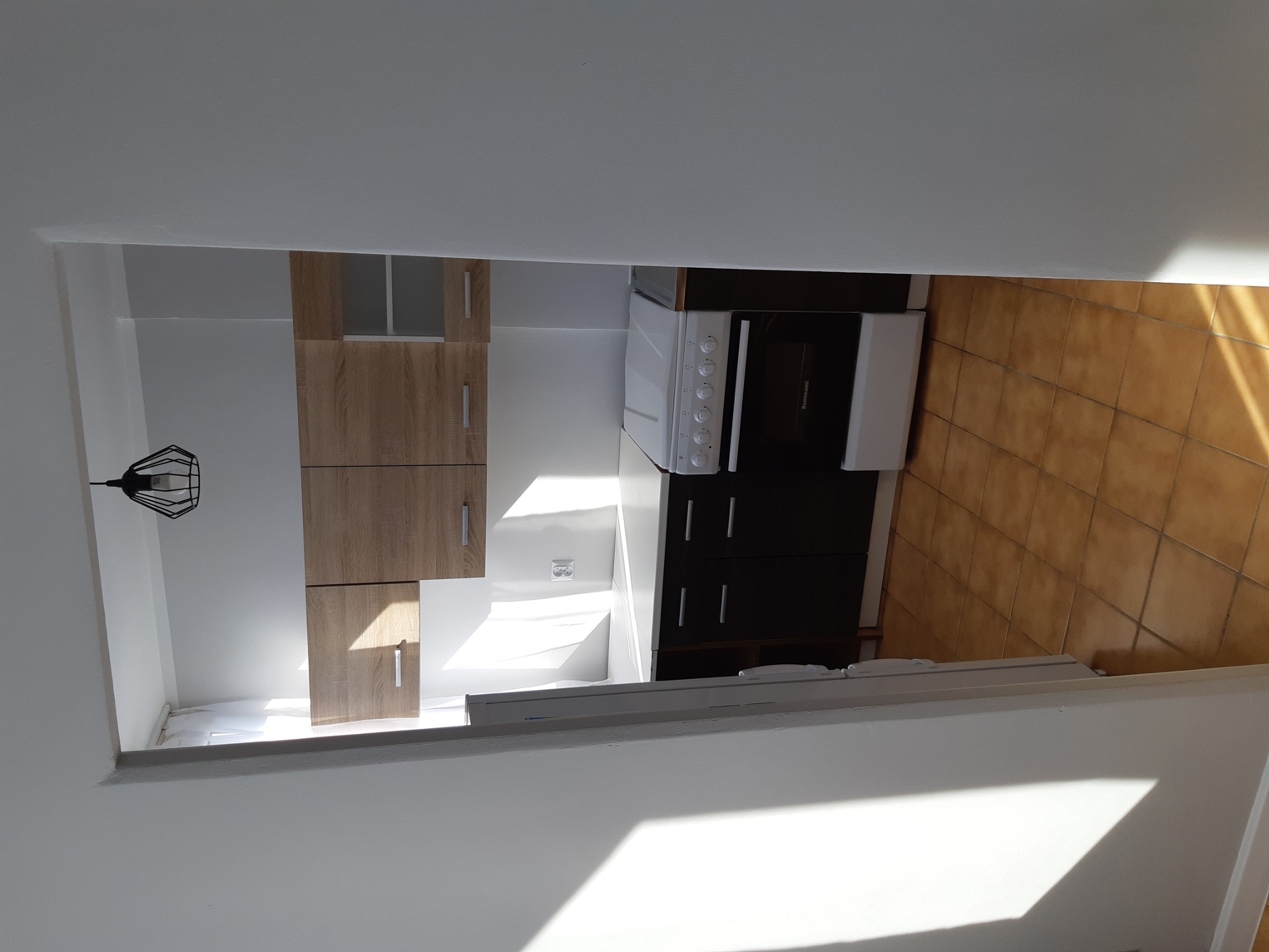 Ukończona kuchnia z drewnianymi szafkami wiszącymi i ciemnymi szafkami dolnymi, białą kuchenką i terakotową podłogą, widziana z góry pod kątem.