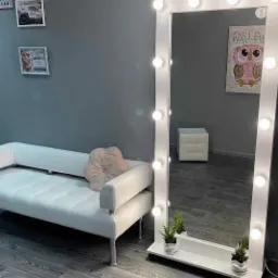 Nowoczesne wnętrze z białym, pikowanym siedziskiem, ozdobną poduszką w kształcie kwiatu i dużym lustrem z żarówkami, doświetlające szarą ścianę z obrazkami i rysunkiem sowy.