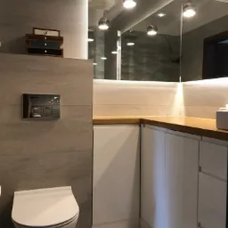 Nowoczesna łazienka z podwieszaną toaletą, szarą płytką imitującą kamień, drewnianym blatem i lustrem z oświetleniem punktowym, w odbiciu widoczna kabina prysznicowa z dekoracyjną ścianą.