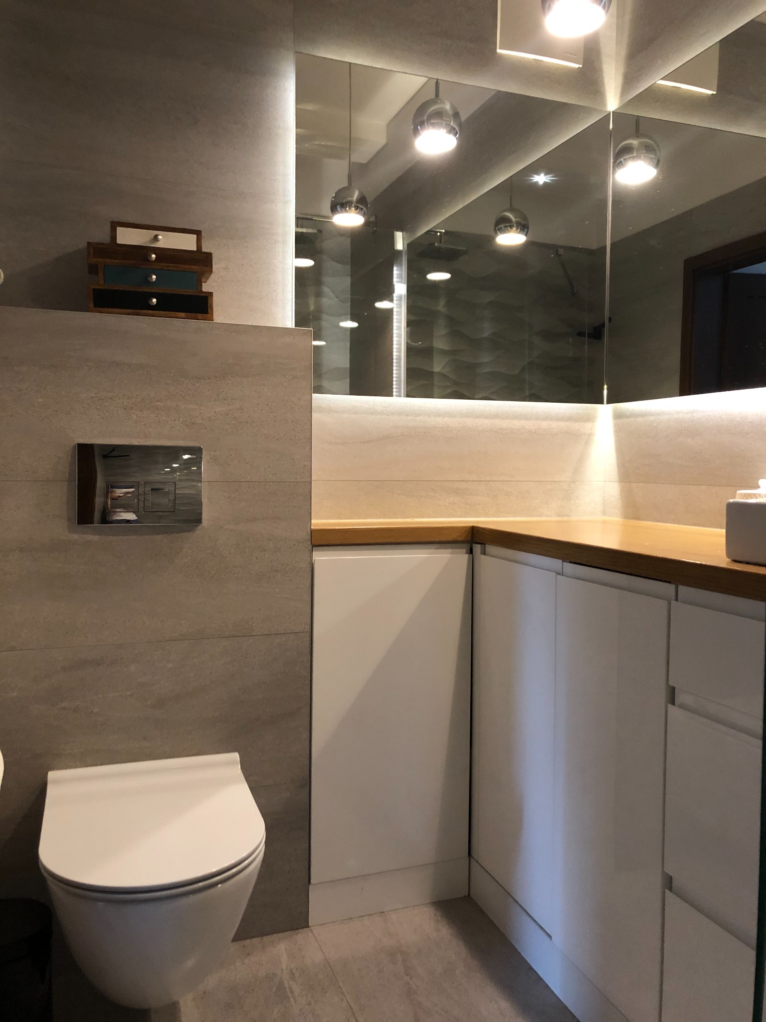 Nowoczesna łazienka z podwieszaną toaletą, szarą płytką imitującą kamień, drewnianym blatem i lustrem z oświetleniem punktowym, w odbiciu widoczna kabina prysznicowa z dekoracyjną ścianą.